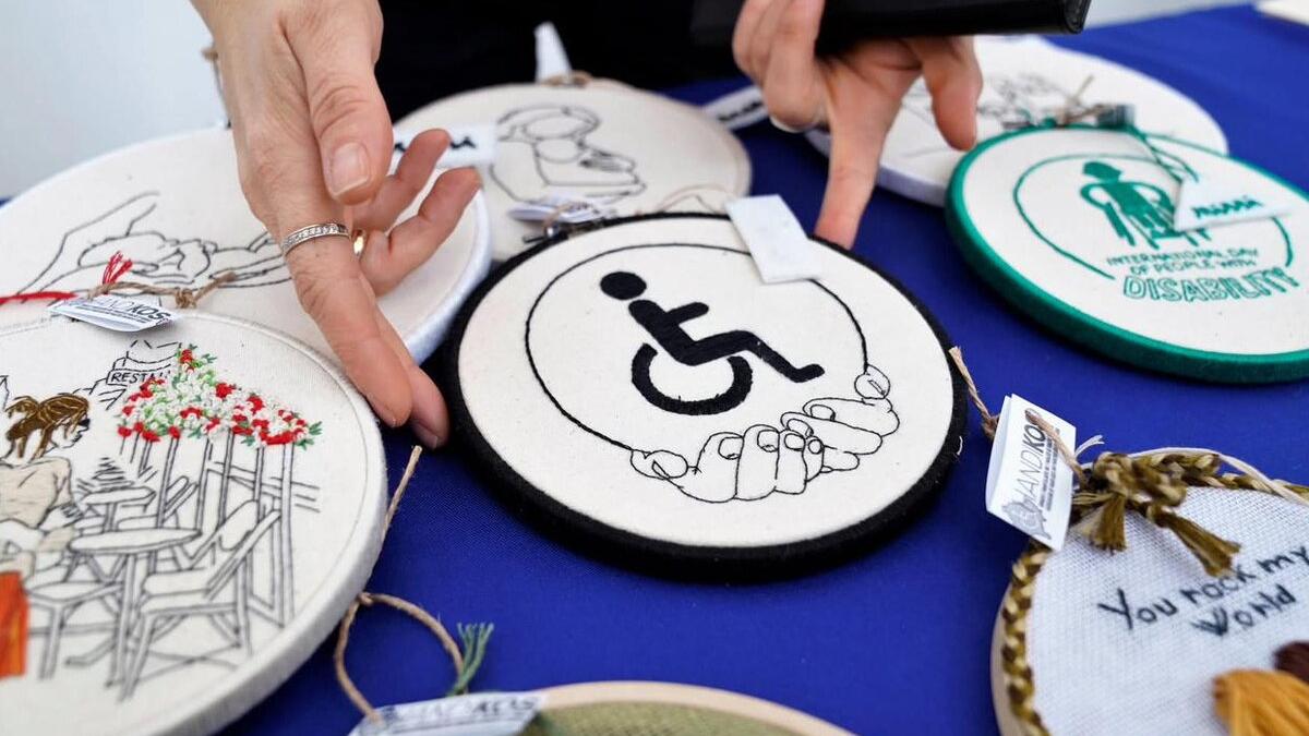Close-up of embroidered circular patches displayed on a blue surface. One patch in the center shows a wheelchair accessibility symbol held by two hands. Other patches feature various designs, including flowers and text.