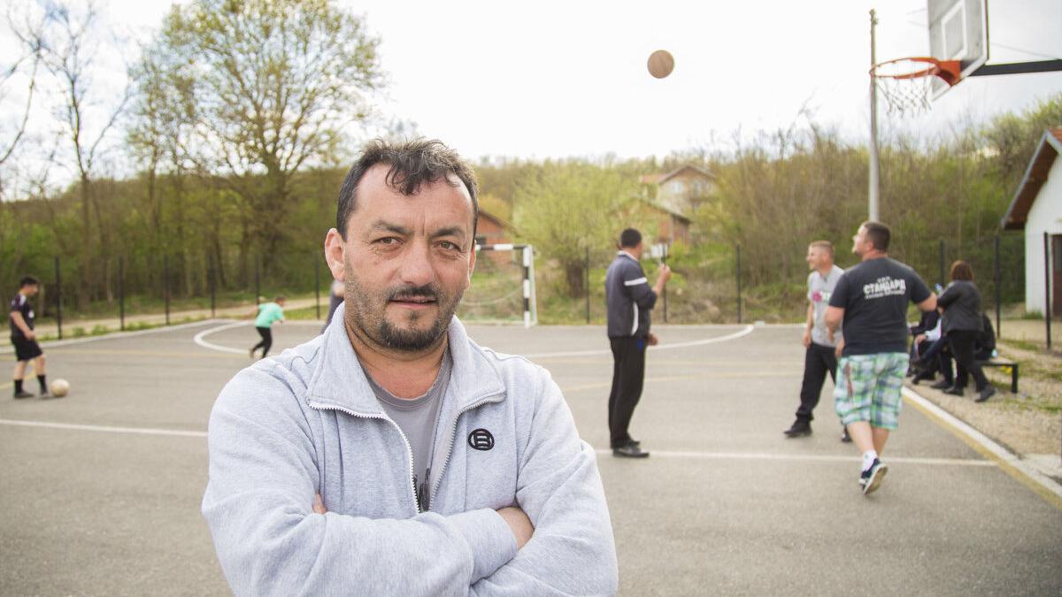 A person stands in the foreground of an outdoor basketball court with arms crossed, wearing a light gray zip-up jacket. In the background, several people are playing or interacting around the basketball court—some dribbling a ball, others talking near the hoop. Trees, small buildings, and a fence surround the court area.