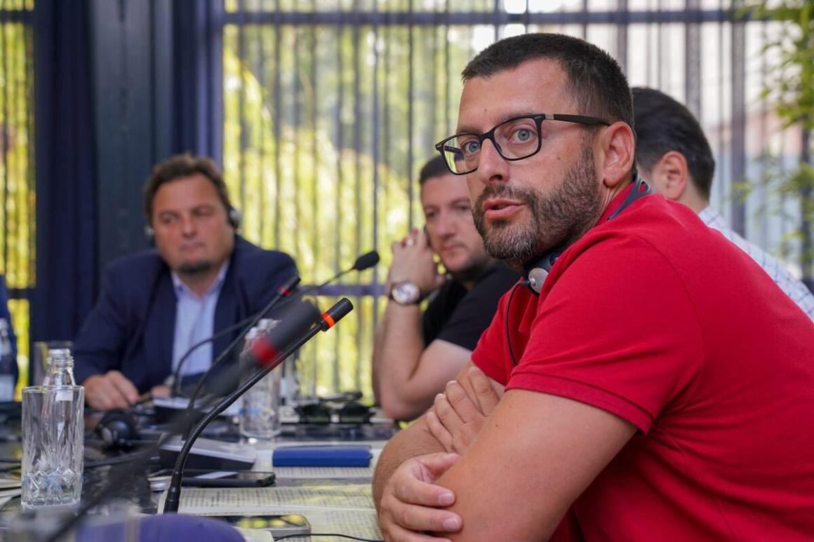 A group of people seated around a conference table with microphones, water bottles, and documents. One person in the foreground is wearing a red shirt and sitting with arms crossed, while others are engaged in discussion. The setting appears to be indoors with large windows and vertical blinds in the background.