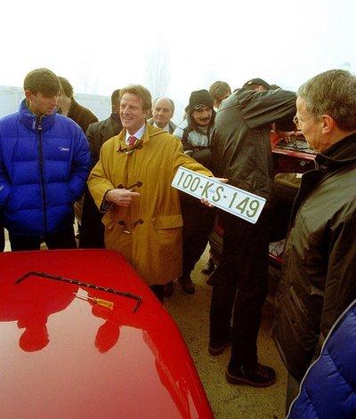 A group of people gather around a red car with its hood open. One person holds a white license plate with black characters reading “100-KS-149,” while others observe closely. The scene appears to take place outdoors in a foggy setting.
