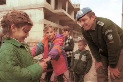 A uniformed peacekeeper wearing a blue beret is leaning forward to speak with a group of children outdoors. The children are dressed in colorful winter jackets and stand near a partially constructed concrete building in the background.